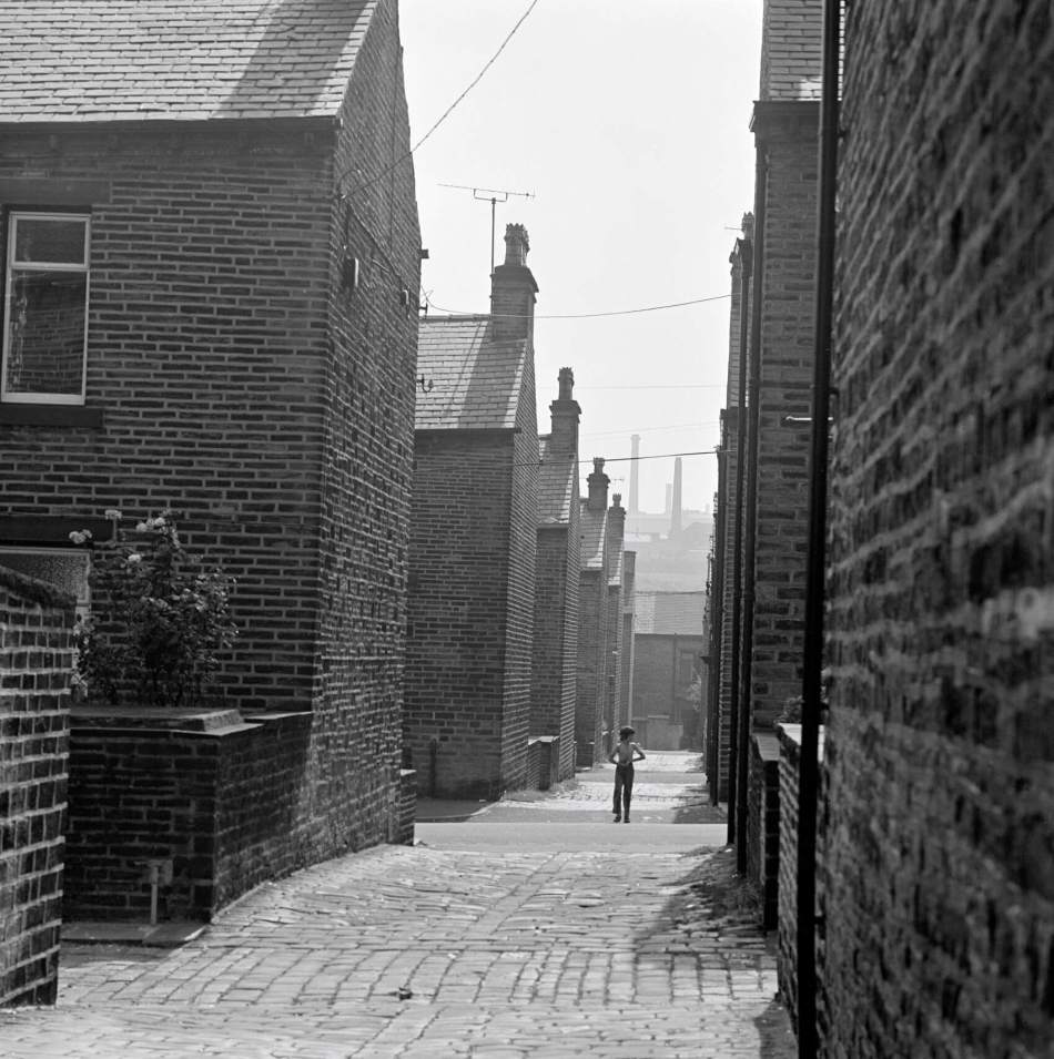 A black and white photograph of a child walking down an alleyway in between rows of terraced housing.