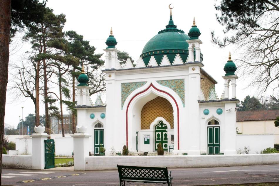 A photograph of the exterior of a mosque with a rendered entrance topped with embattlements and open turrets, surmounted by a large dome.
