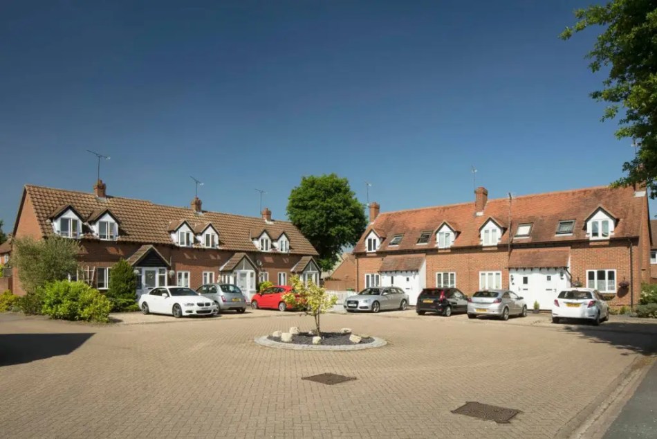 A photograph of 2 small terraces of houses with cars parked outside the entrances beside a large paved area with a small tree in the middle.