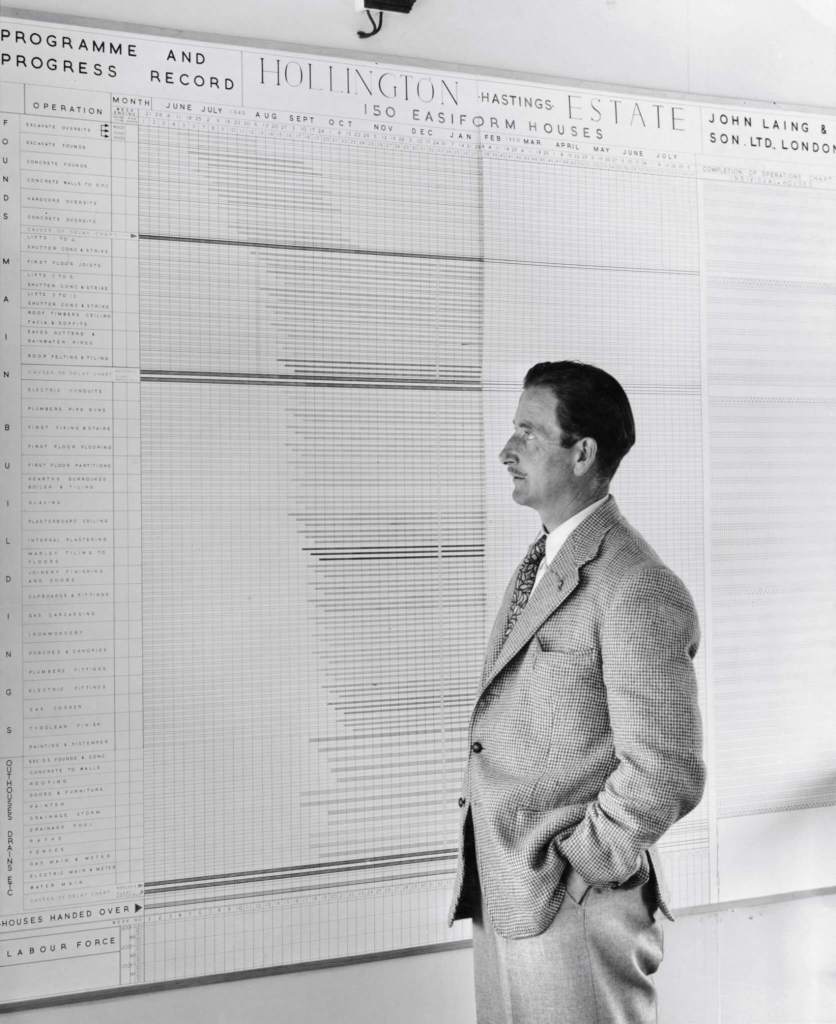 A black and white photograph of a man wearing a suit standing in front of a board recording the progress of housing estates building.