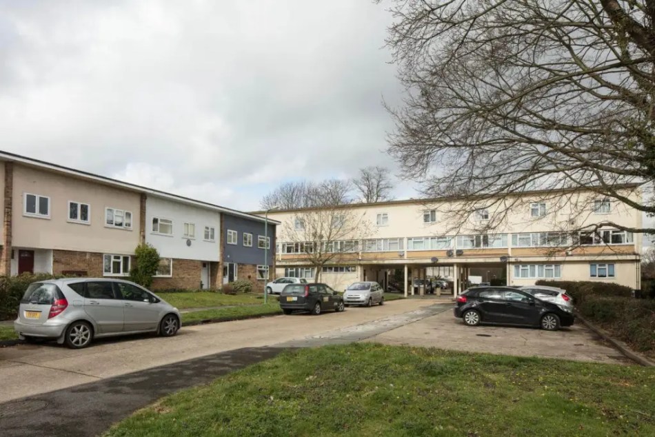 A photograph of a row of terraced houses beside a small block of flats beside an access road. Cars are parked on the road and in parking spaces in front of the buildings.