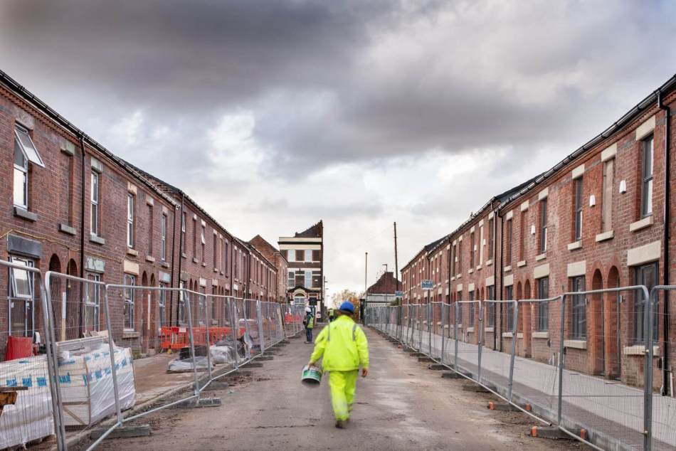 A photograph of a person wearing high visibility trousers and a jacket walking down the middle of a road with terraced housing on either side. Temporary metal gates stand in front of the entrances to the houses as well as some building materials.