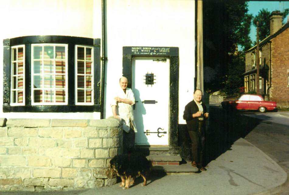A photograph of two men and a dog stood outside a house. 