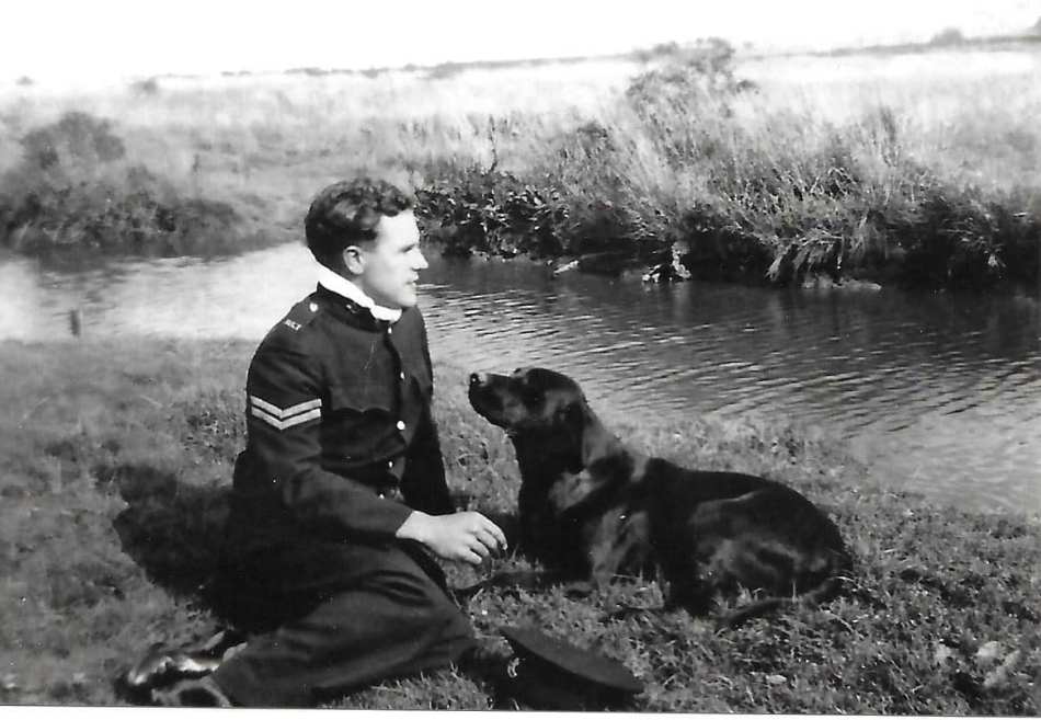 A photograph of a man in uniform sat by a lake with his dog. 