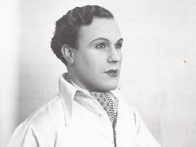 A black and white portrait photograph of a man with pencil eyebrows and lipstick. 