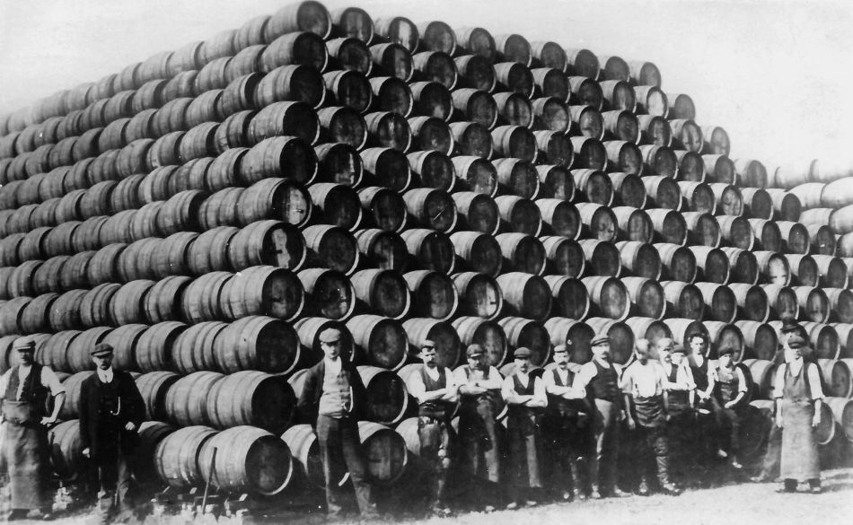 A black and white photograph of coopers standing in front of an enormous pyramid of barrels.