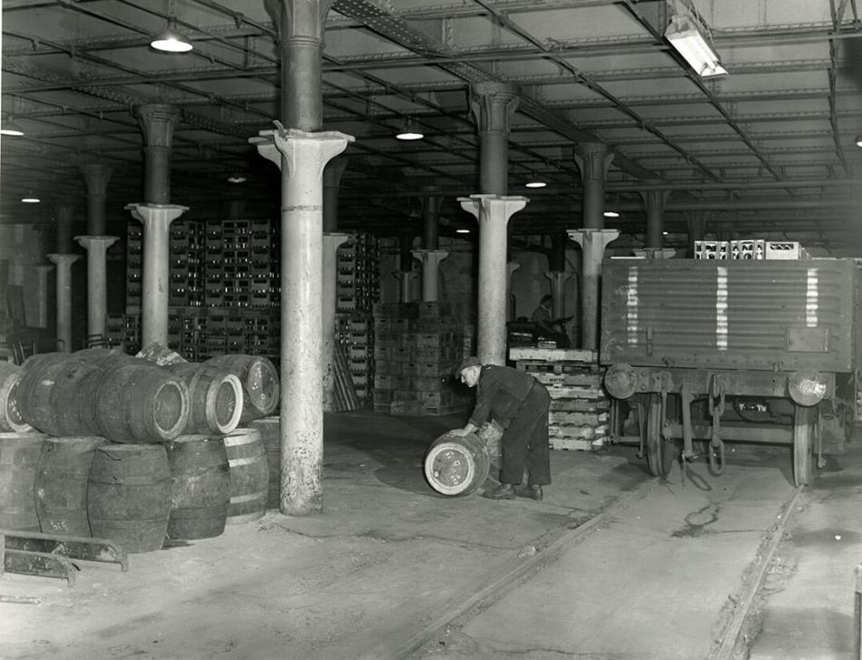 A black and white photograph of an undercroft filled with barrels. A person rolls one barrel.