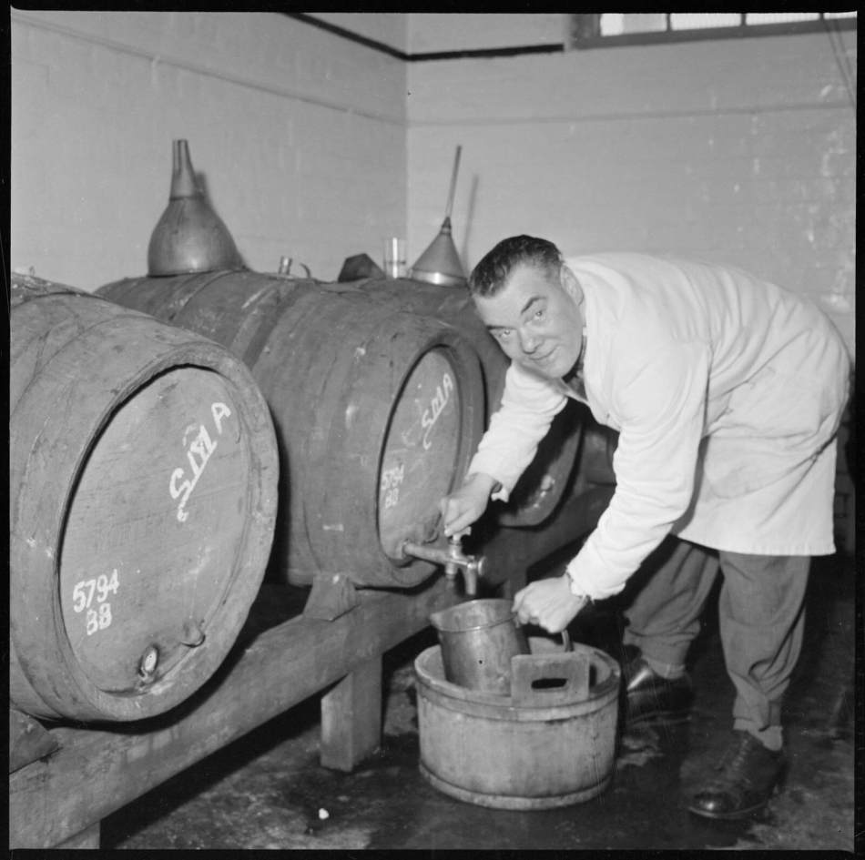 A black and white photograph of a man in a white coat bending over to drain beer from a barrel tap while looking at the camera.