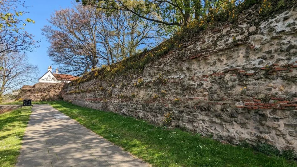 A photograph of the remains of a historic town wall beside a modern pavement.