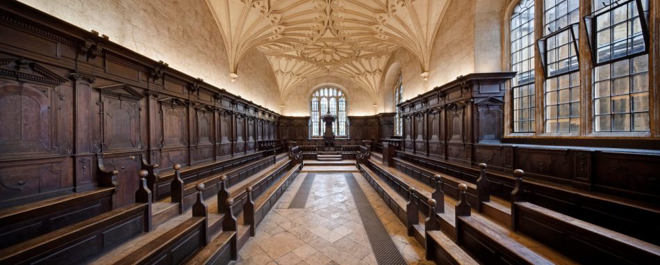 A photograph of the interior of a long room with wooden panelling on the walls and pews on either side of the room. The ceiling features decorative fan vaulting.