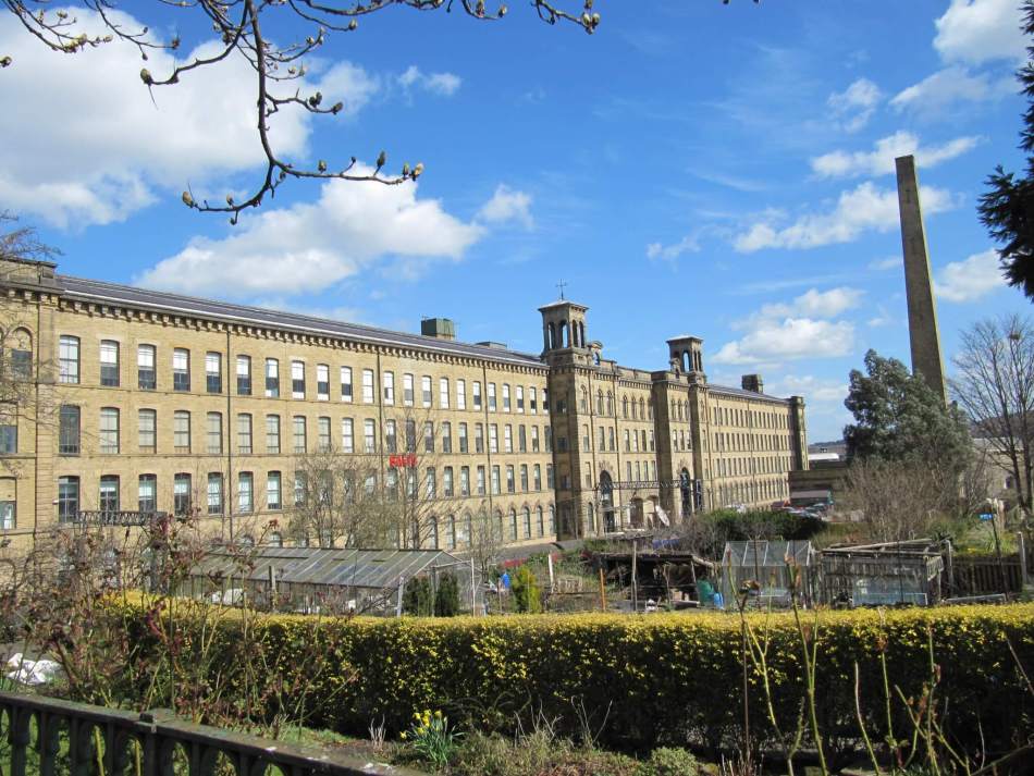 A photograph of the exterior of a large mill building surrounded by allotments and greenhouses.