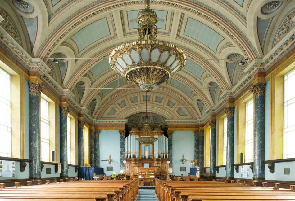 A photograph of the interior of a church with ornate ceiling decoration and large elegant light fittings hanging above the centre of the aisle.