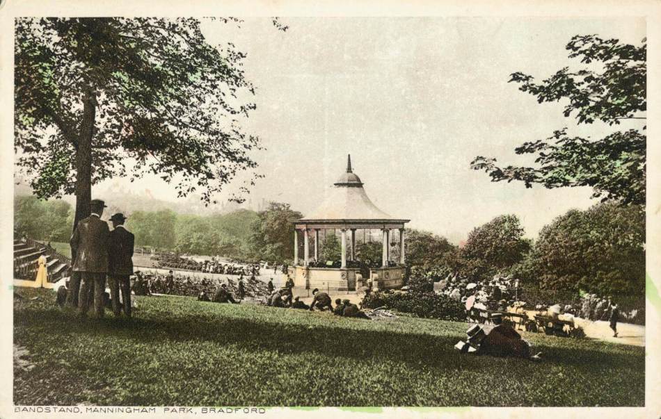 A scan of a postcard featuring a photograph of a bandstand in the centre of a park with people sat on the grass surrounding it.