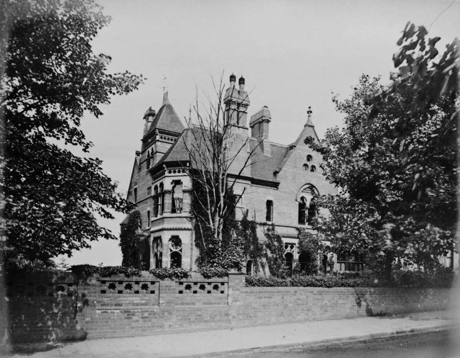 A black and white photograph of a Gothic style villa surrounded by trees and a brick wall.
