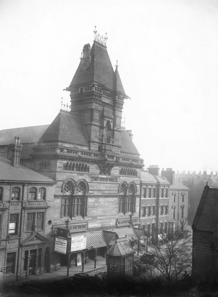 A black and white photograph of a tall music hall with a small cabmen's shelter on the road beside the building.