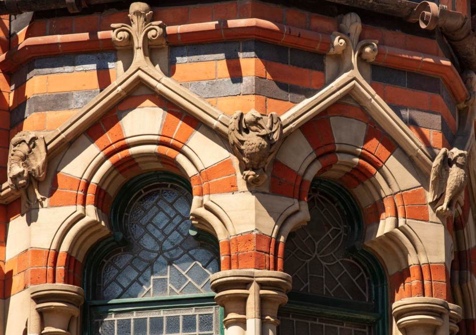 A photograph of a close-up of carved gables above trefoil-headed windows on the projecting turret on an ornate red brick building.