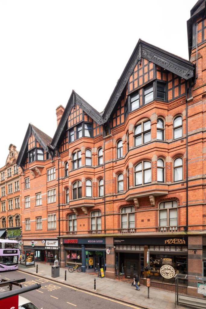A photograph of a terrace of 5-storey large red brick buildings with shops on the ground floor. The upper storeys have timber-framed gables, dormers and elegant windows.