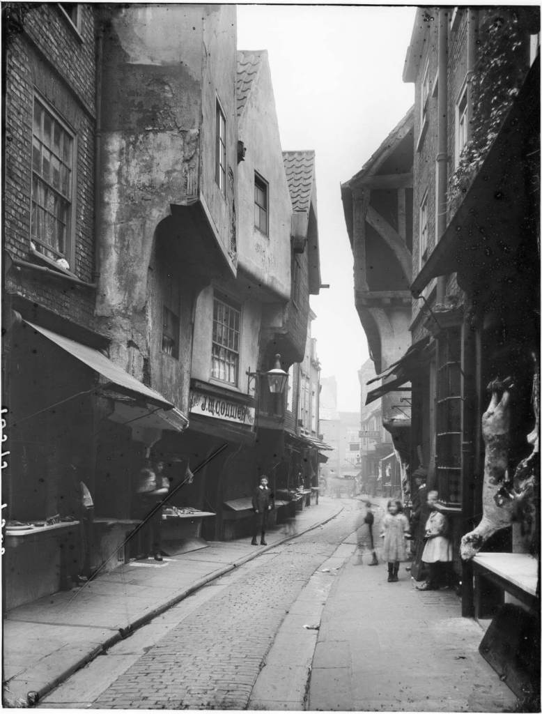 A black and white photograph of a high street with medieval buildings. Animal carcases are hanging from the windows of butchers shops. 