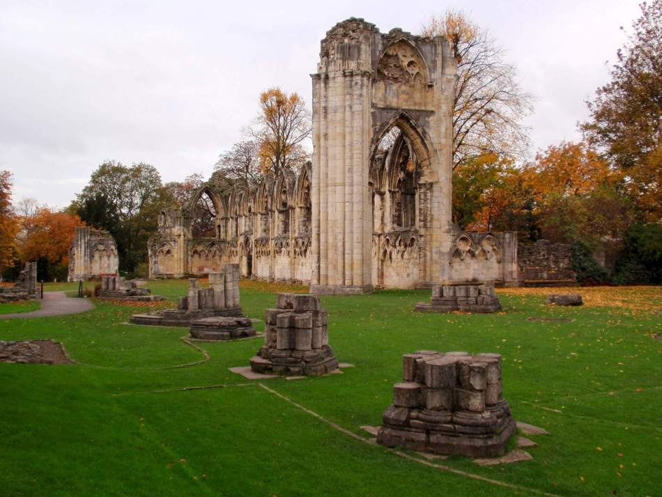 A photograph of church ruins surrounded by autumnal trees, with small column stumps surrounded by grass in the foreground
