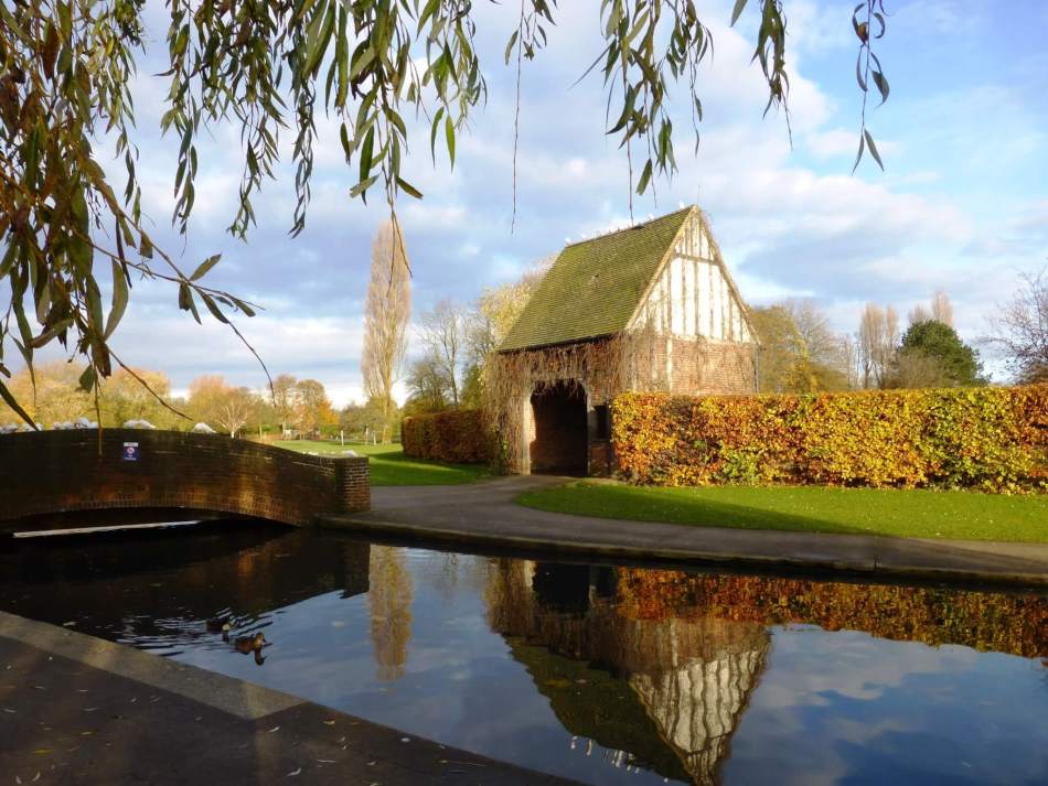 A photograph of a park scene featuring an arch-roof building, hedging, and a waterway and bridge in the foreground.