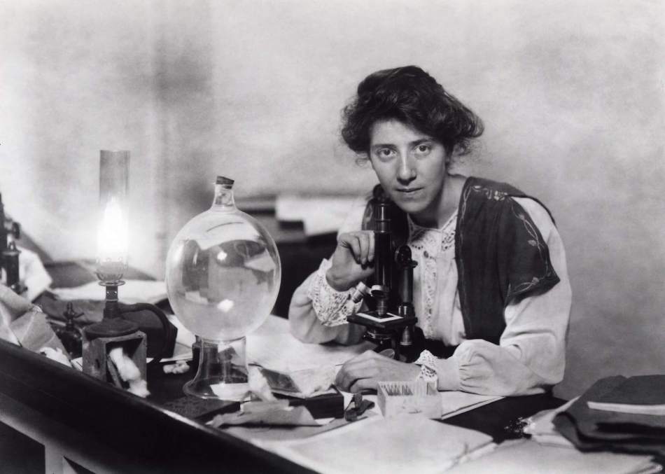 A black and white photograph of a woman sat at a desk leaning over a microscope
