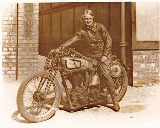 A sepia photograph o a woman wearing motorcycle leathers sat on top of a motorcycle. 