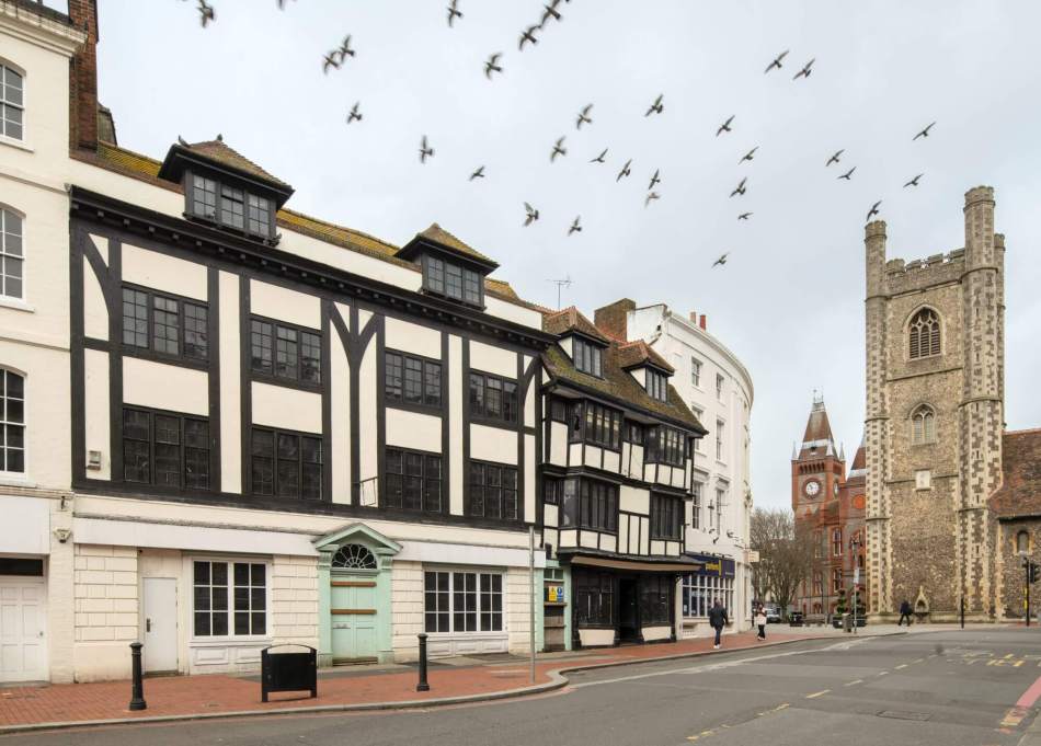 A photograph of timber-framed buildings beside a road with a church tower in the background.