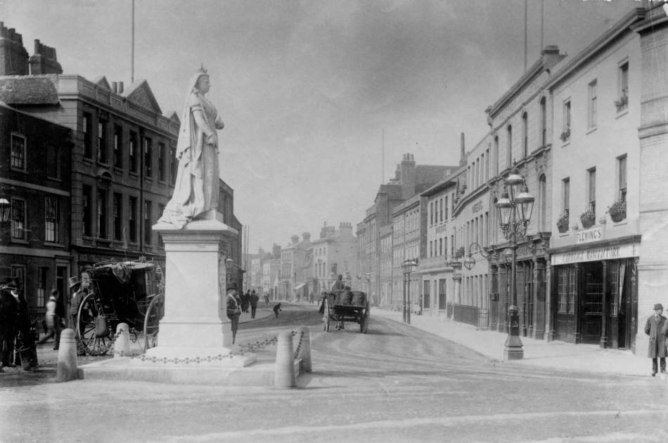 A black and white photograph of a tall statue of Queen Victoria with a high street in the background.