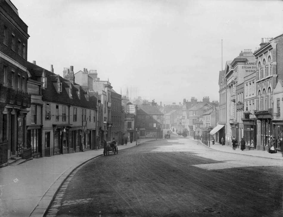 A black and white photograph of a high street with a person travelling in a small horse and carriage down the road.