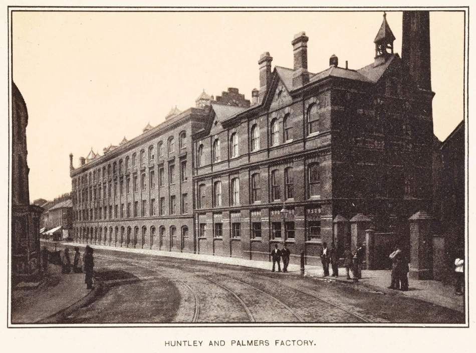 A black and white photograph of the exterior of a large factory building beside a road.