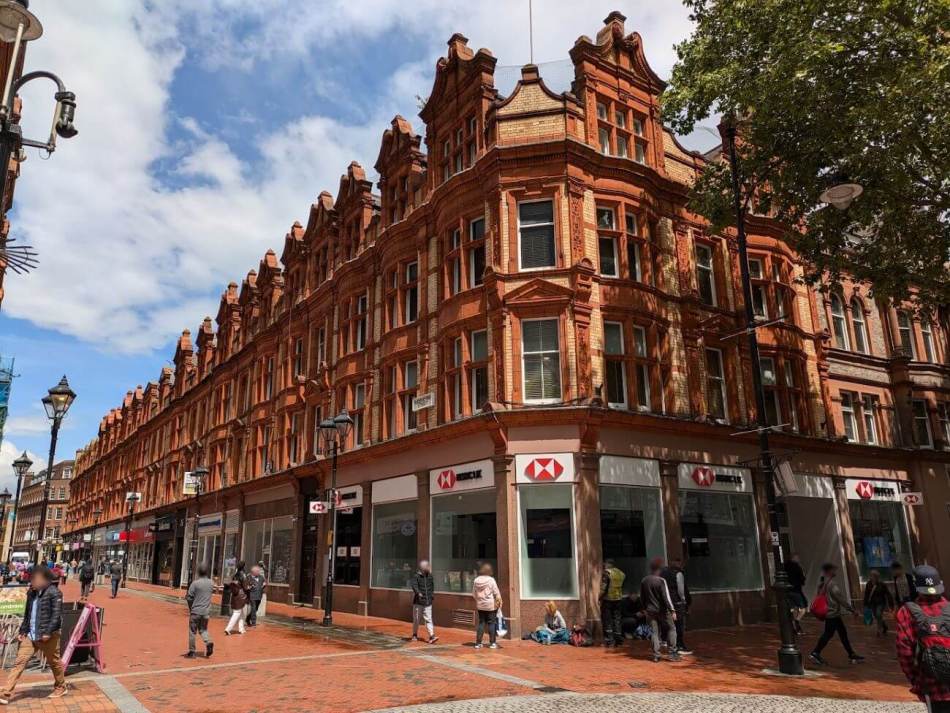 A photograph of the corner of a busy high street with shops on the ground floor and 3 storeys of ornate red brick building above.
