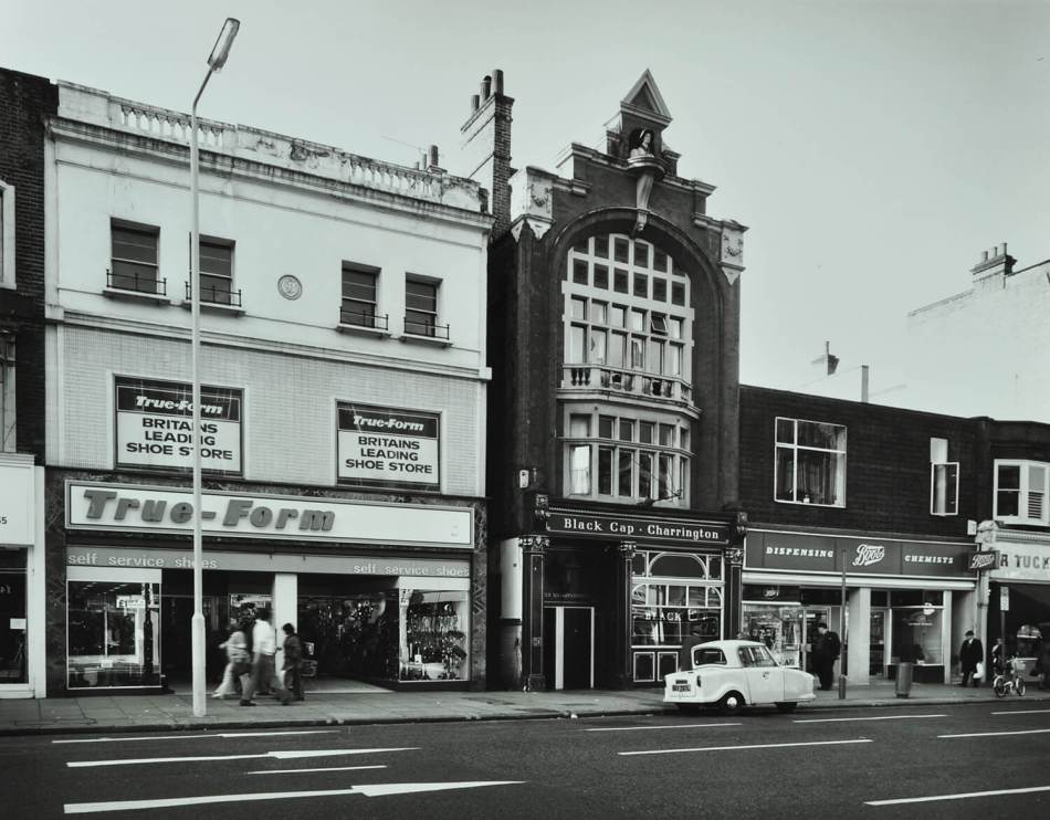 A black and white photograph of a high street with a row of shops and a pub.