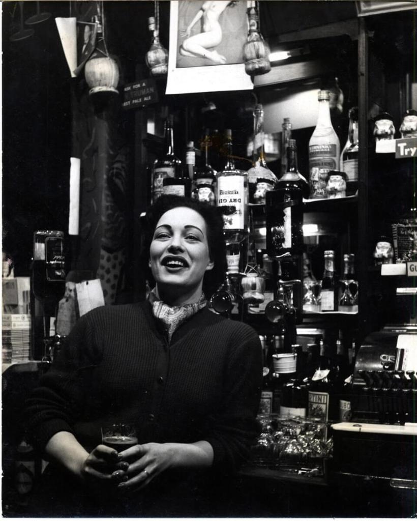 A black and white photograph of a woman holding a drink, leaning against a bar with lots of liquor bottles on it.