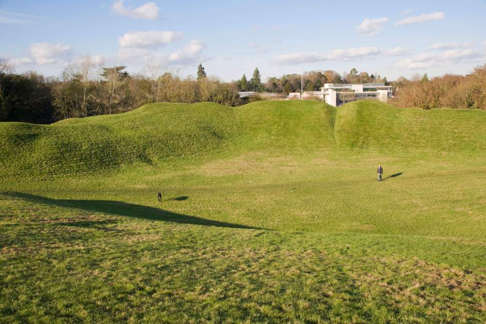 Photograph of the earthwork remains of an amphitheatre, the small figures of visitors  give an idea of the height of the earthwork  banks where tiers of seating once were.