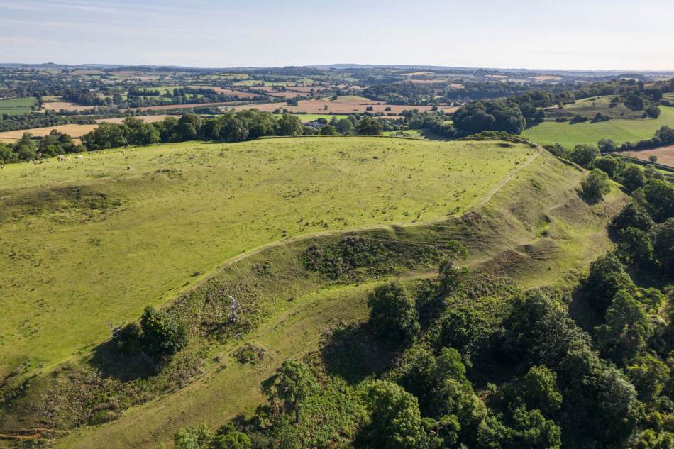 An aerial view of a hill fort with multiple banks, the lower slopes are tree-grown.