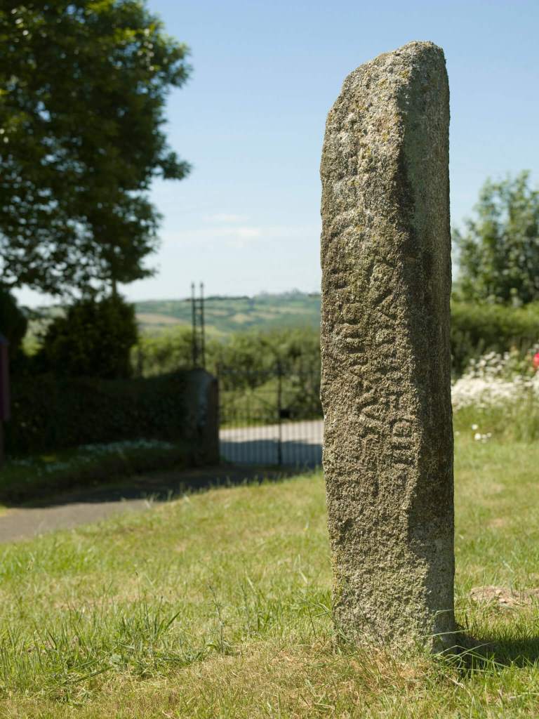 A photograph of a weathered stone pillar in a churchyard.