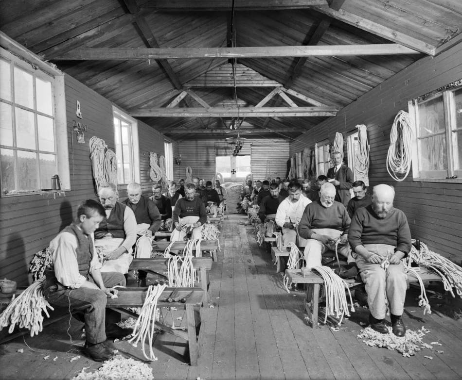 A black and white photograph of rows of men and a boy sitting on benches in a hut, making rope handles.