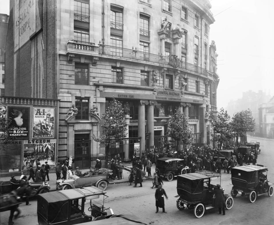 A black and white photograph of a crowd of people and a busy road of old cars.