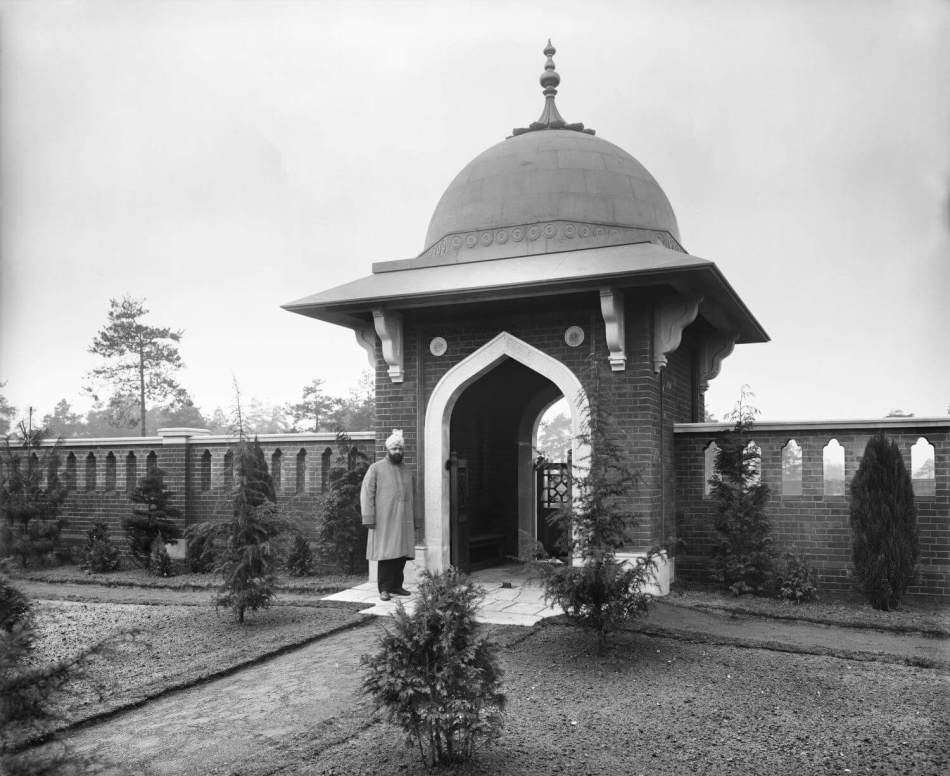 A black and white photograph of a man standing in front of the entrance to a burial ground.