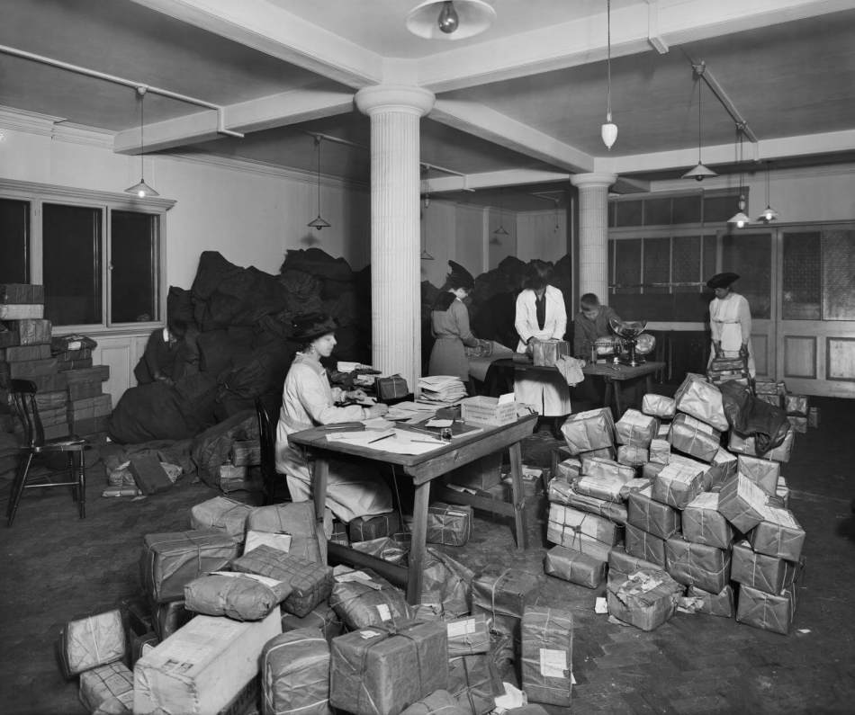 A black and white photograph of women working through piles of packages in a postroom.
