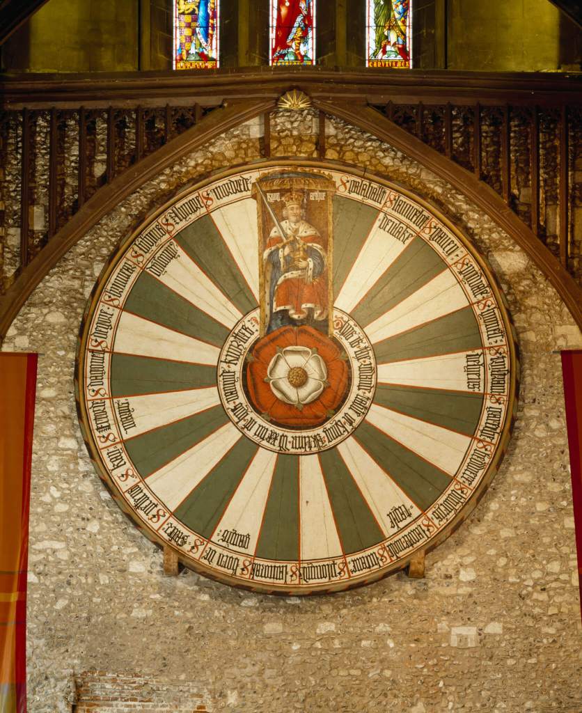 A huge wooden round table top painted and embellished with the heraldic motifs and gothic script, set on an internal stone wall of a building.