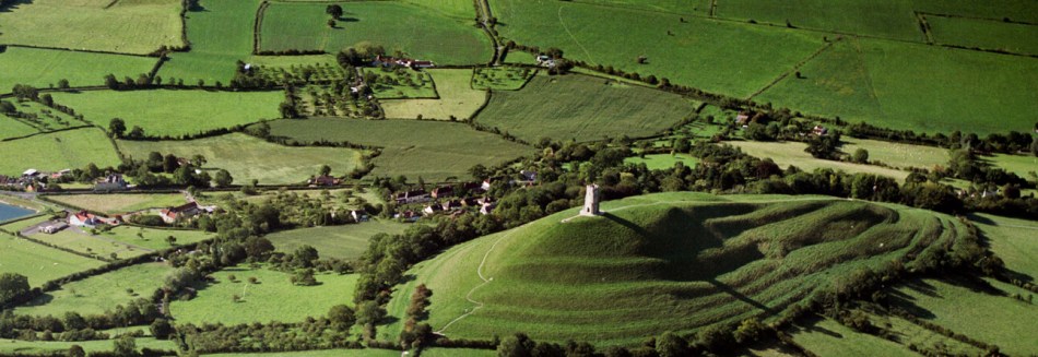 An aerial view of a hill surmounted by a tower set in an otherwise flat landscape.