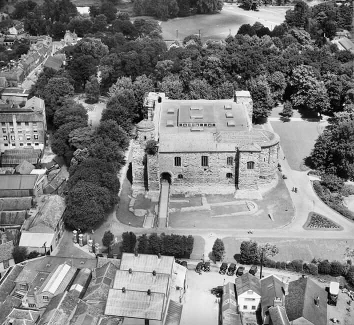 A black and white photograph of a brick two storey castle surrounded by trees. 
