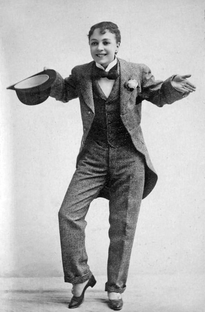 A black and white photograph of a woman in a suit with a top hat, smiling at the camera. 