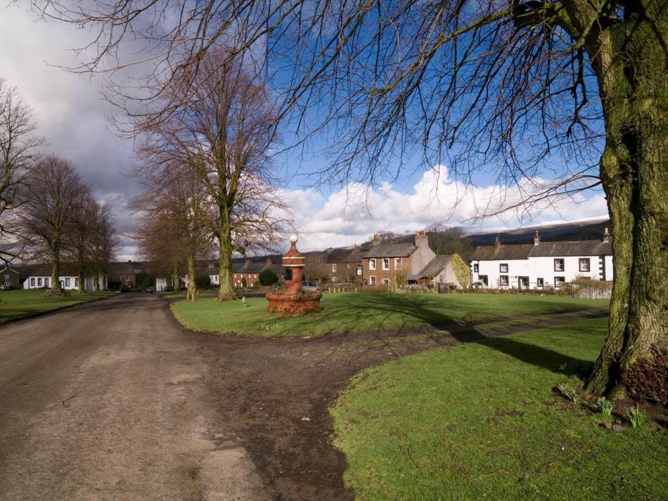 A photograph of a village green, surrounded by cottages, with a large water pump in the middle of the green.