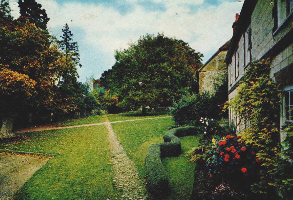 A photograph of a postcard showing a village green surrounded by trees beside a cottage.