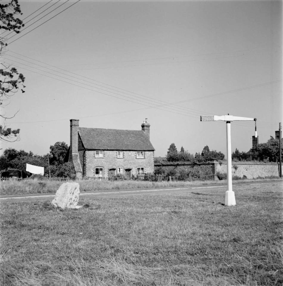 A black and white photograph of a tilting pole in a middle of a village green, with 2 cottages in the background.