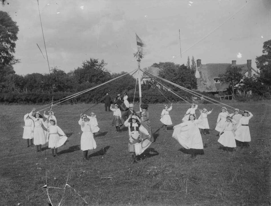 A black and white photograph of children dancing around a maypole on a village green.