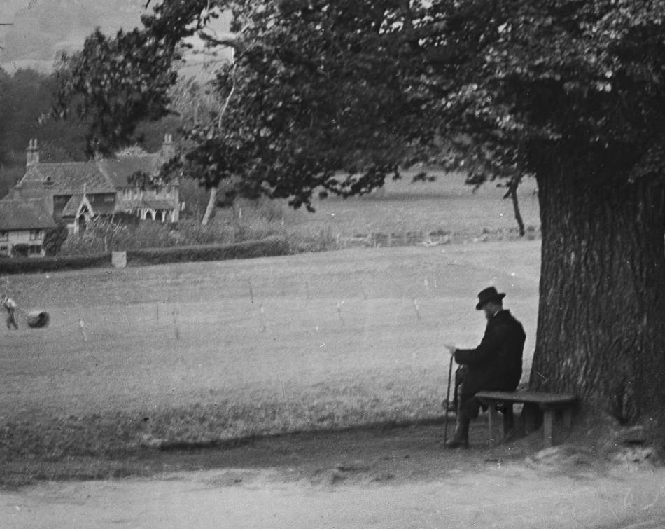 A black and white photograph of a man sitting on a bench in front of a tree, overlooking a village green.