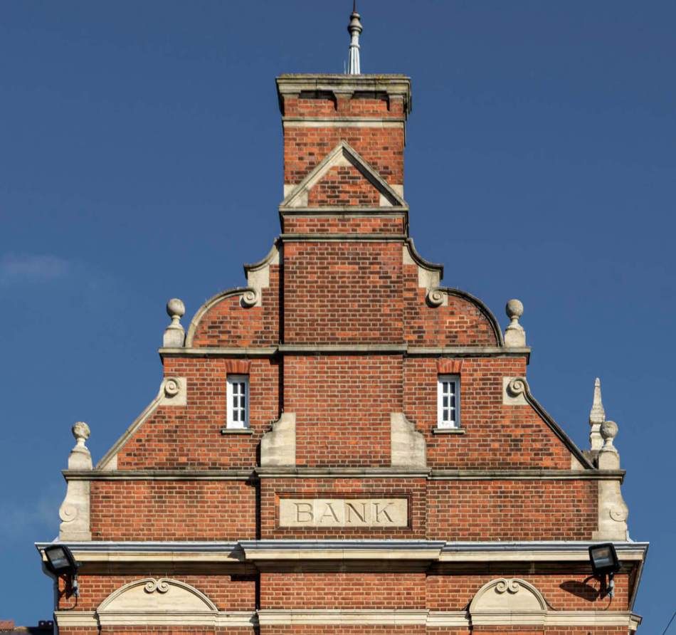 A close-up photograph of a red brick bank building with a carved stone reading 'BANK'.